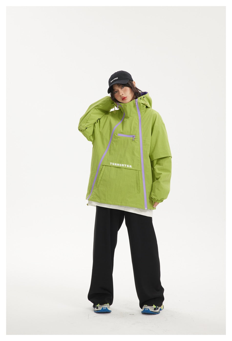 Model in lime green hooded jacket with lilac zippers, hand raised to head, black cap and black wide-leg pants