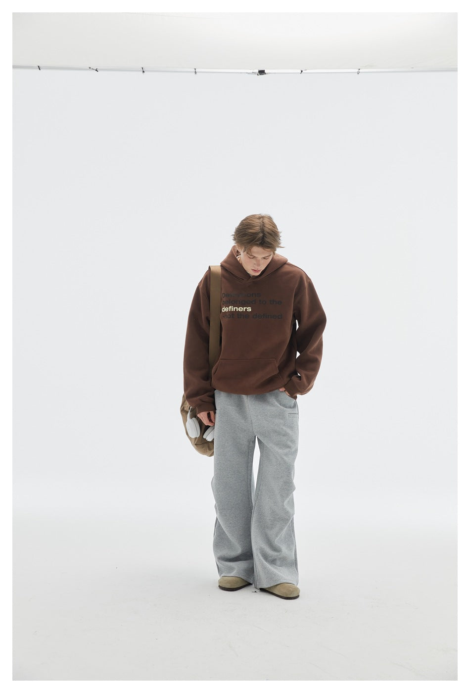 Front view of a model wearing a brown text-print hoodie and wide gray pants, carrying a shoulder bag and headphones.