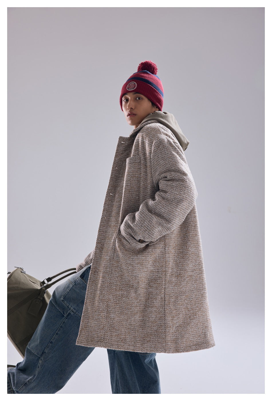 Model wearing a long beige herringbone coat with a gray hood, shown in a three-quarter front view.