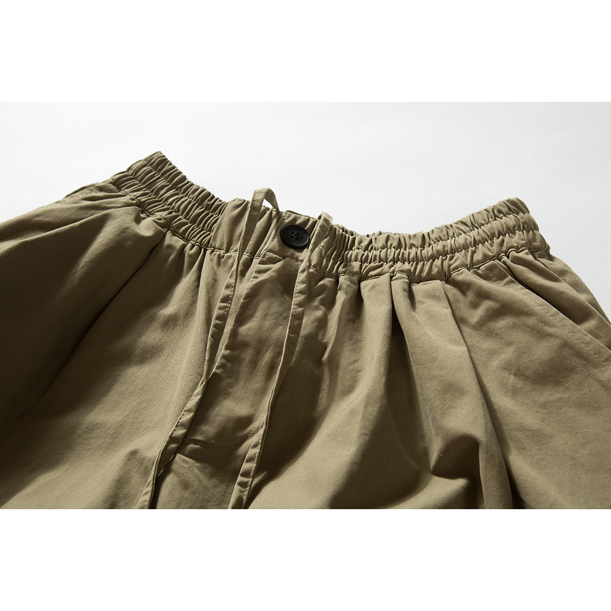 Close-up of khaki trousers showing the gathered elastic waistband, button closure, fly, and front pleats.