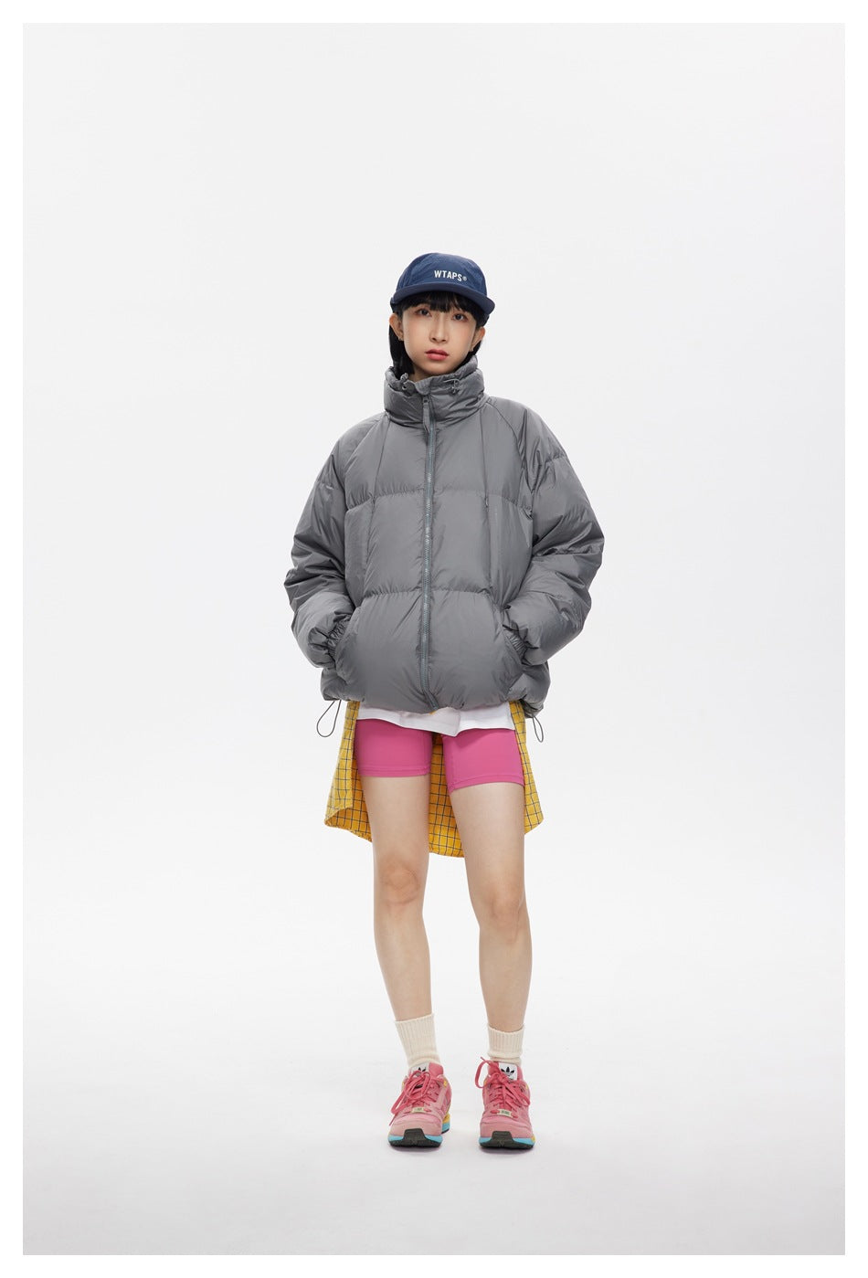 Model wearing a gray high-collar puffer jacket with pink shorts and a yellow checked shirt tied at the waist.