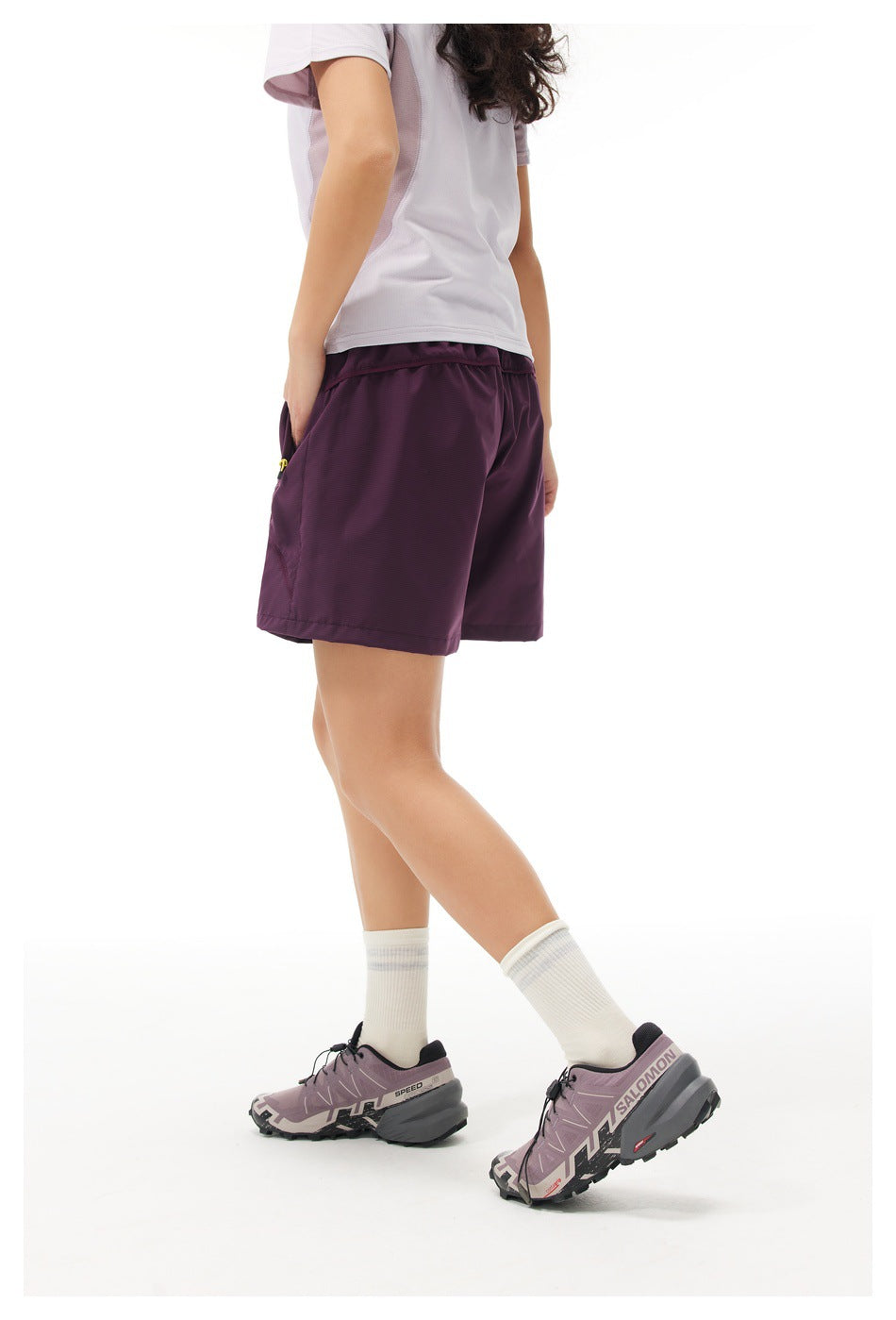 Rear three-quarter view of a model wearing plum athletic shorts with a waistband and side pocket.