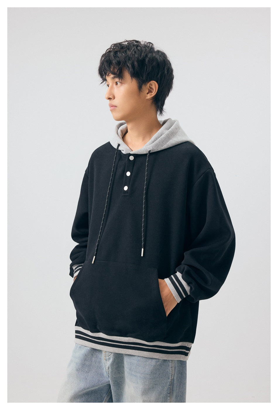 Model wearing black hoodie with gray hood and striped trim, hand in pocket, with light-wash jeans