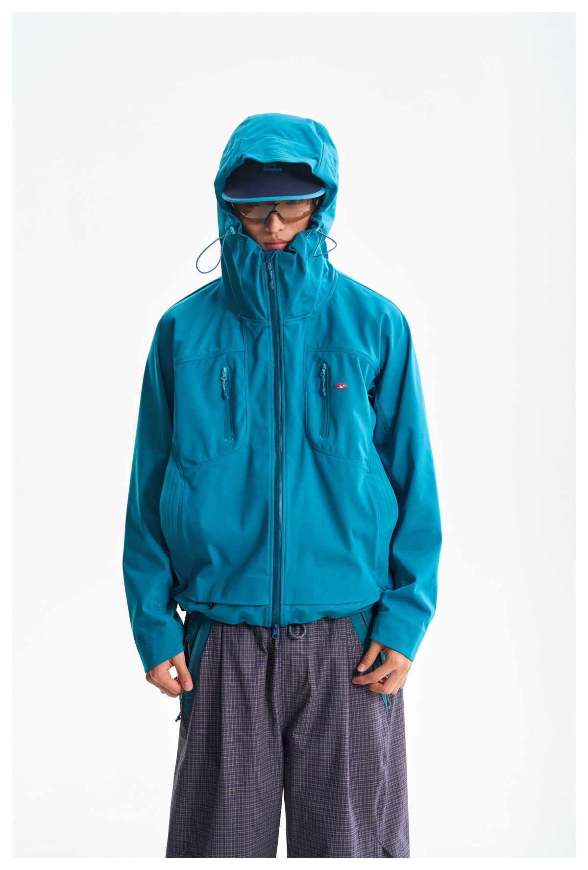 Closer front view of a model wearing the teal jacket with its hood up and two zip chest pockets.
