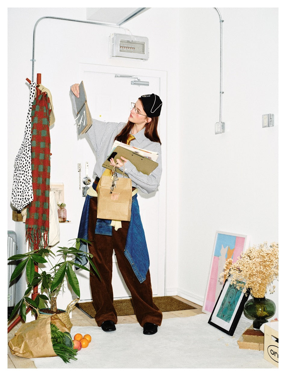 Full-length model in a gray-and-white check shirt, brown trousers, and blue denim tied at the waist, holding papers.