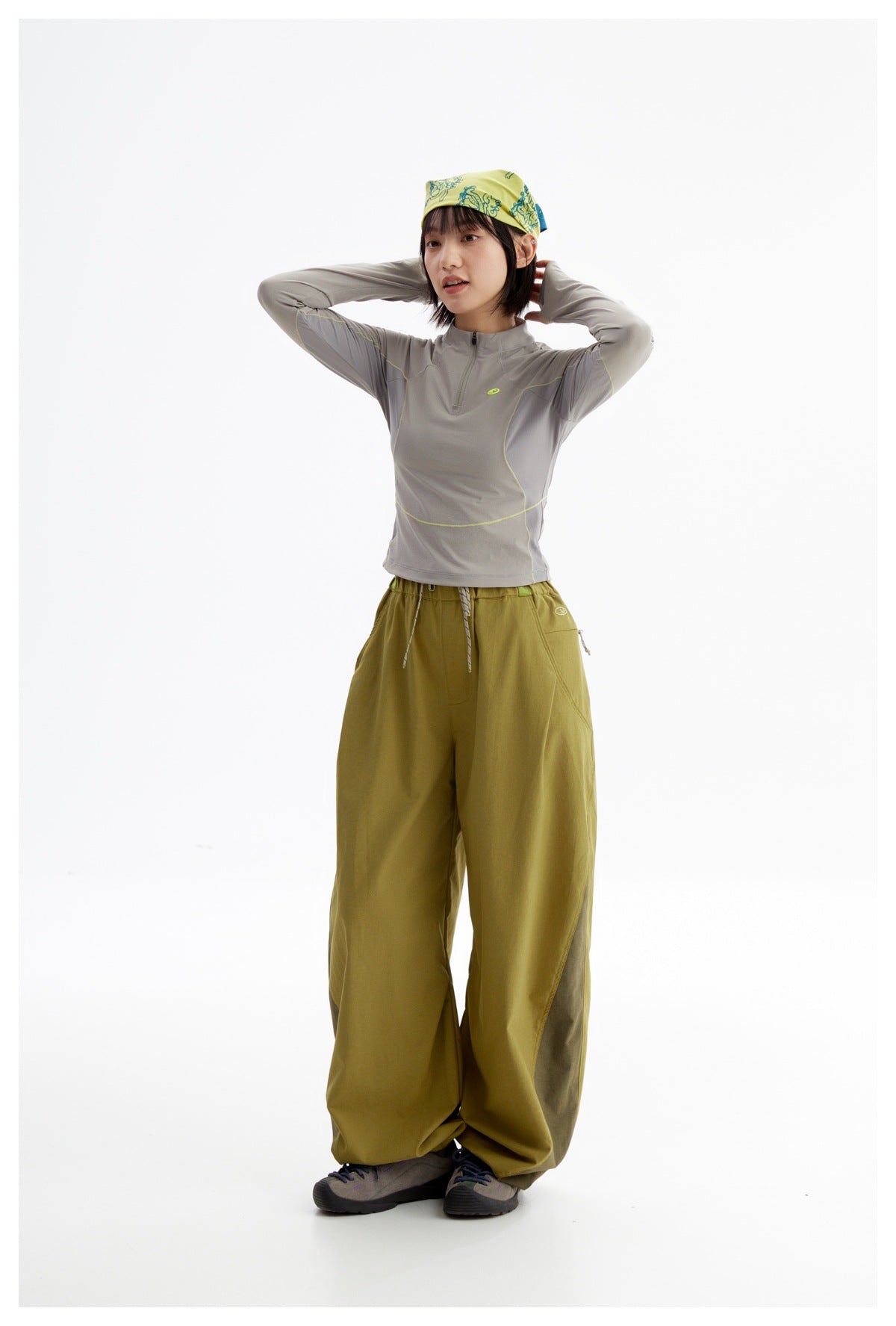Model posing with hands behind her head in a gray quarter-zip top, olive wide-leg pants, and patterned headscarf.