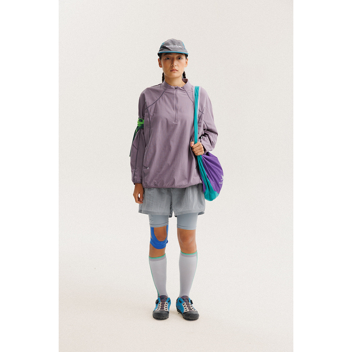 Model wearing a muted lavender quarter-zip pullover with gray shorts and a cap, shown full length.
