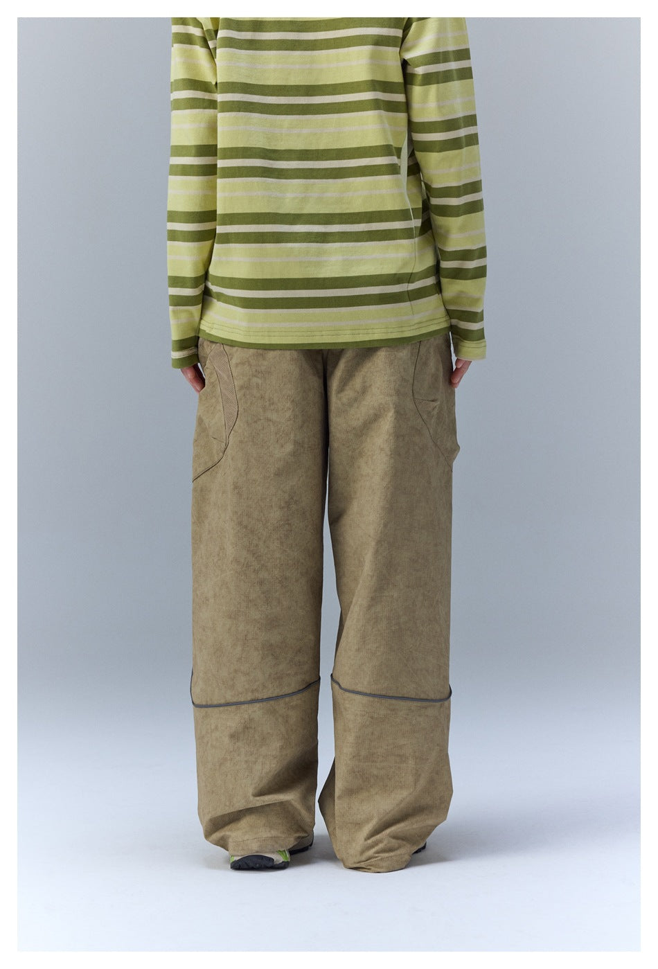 Back view of a model wearing loose khaki pants with cargo pocket panels and horizontal piping at the knees.