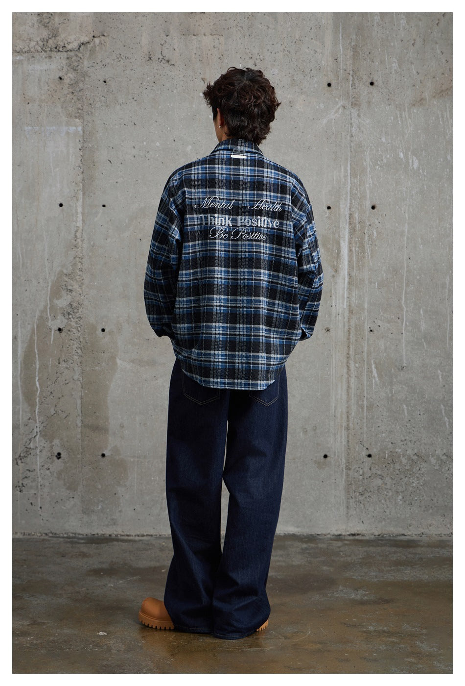 Back view of model in blue plaid shirt with embroidered lettering, dark wide jeans and tan boots