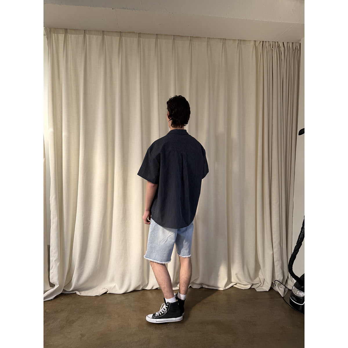 Back view of a model wearing a dark pinstriped short-sleeve shirt and light blue frayed denim shorts.