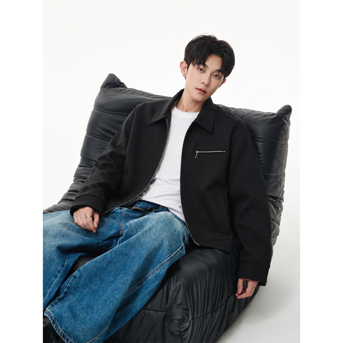 Seated model wearing a black zip-front jacket with a pointed collar and horizontal chest zipper pocket.