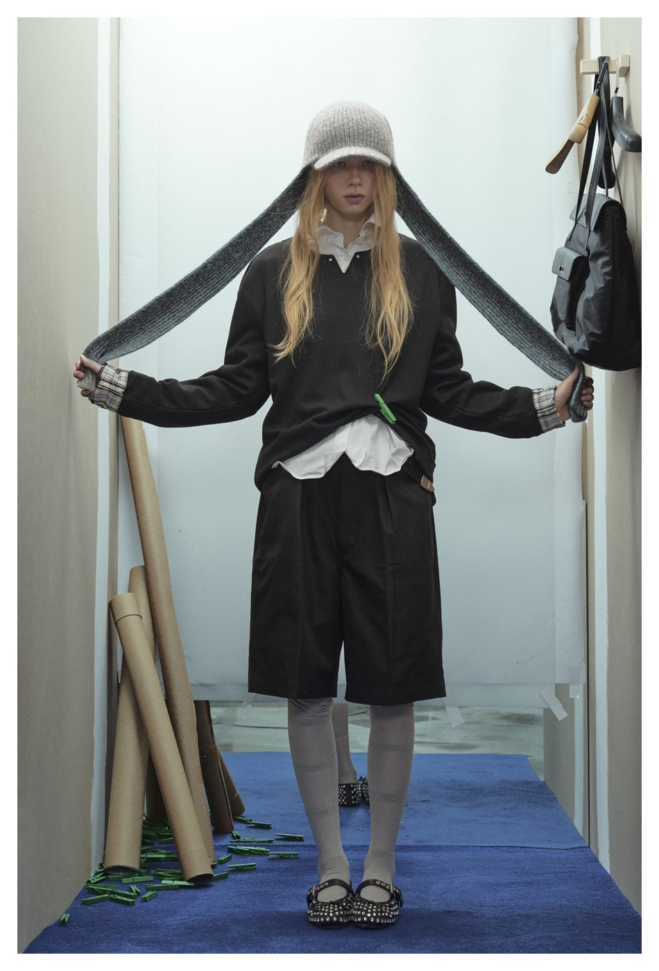 Model wearing a black sweatshirt with plaid cuffs and a white shirt, holding out the long panels of a gray knit cap.
