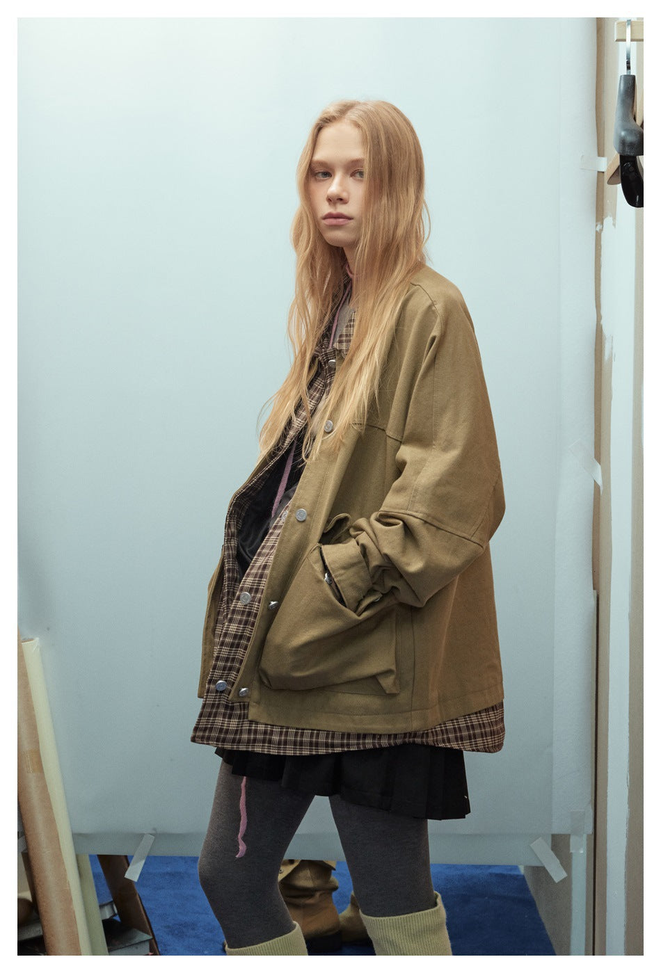 Model wearing an oversized olive-brown jacket with large buttoned pockets over a brown plaid shirt.