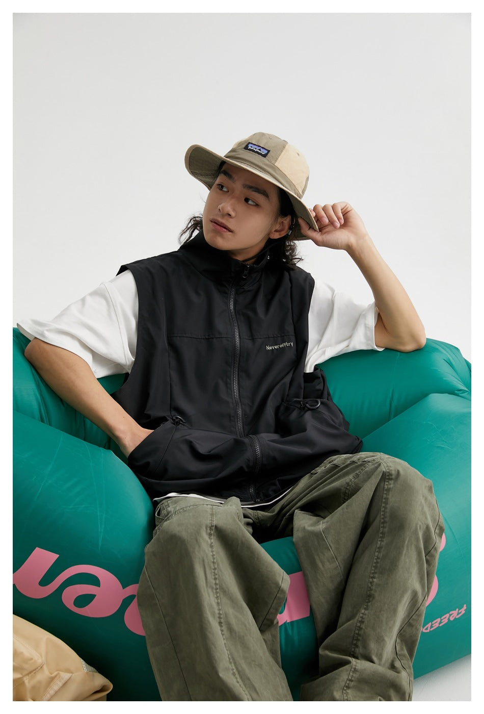 Model seated in a teal chair wearing a black zip-front vest over a white T-shirt and olive trousers.