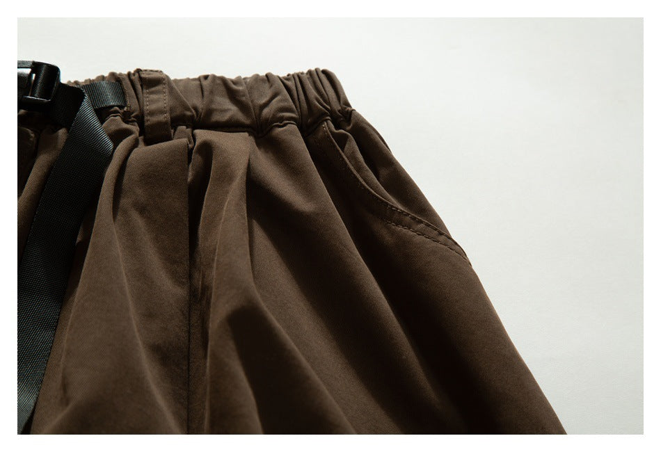 Close-up of dark brown trousers showing the gathered waistband, belt strap, and angled side pocket.
