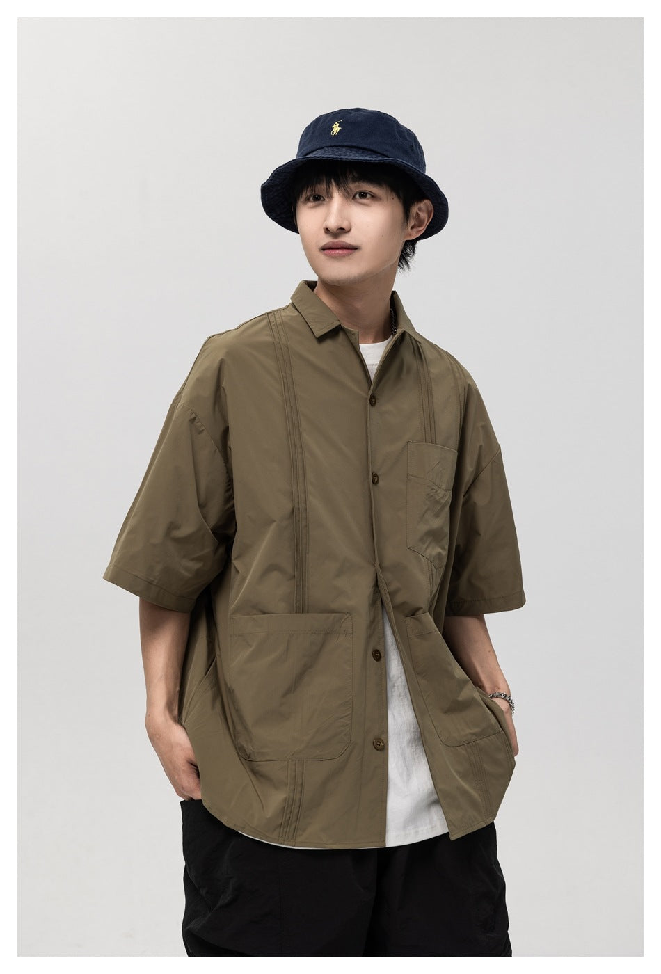 Model wearing an olive-brown short-sleeve button-up shirt with a chest pocket and two large lower pockets.