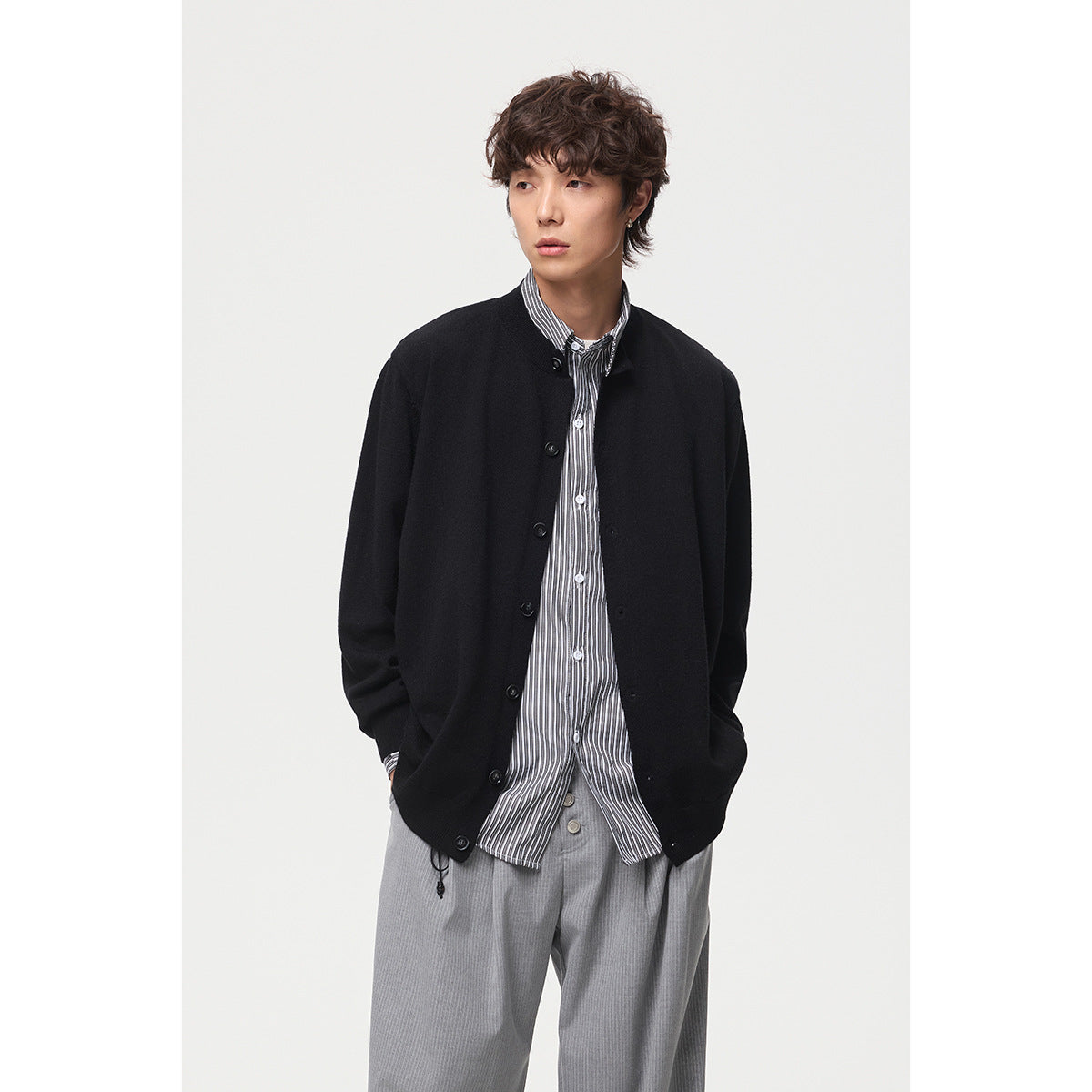 Model wearing a black button-front cardigan over a striped shirt and gray trousers, shown from the front.