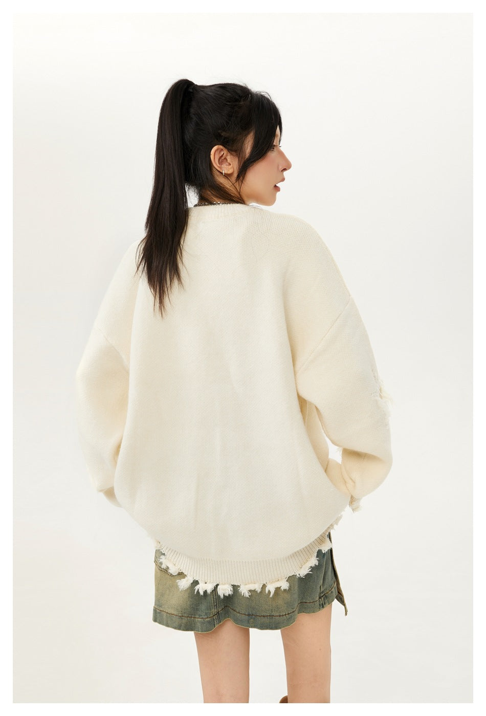 Rear view of a model wearing an oversized ivory sweater with distressed fringe at the hem and a faded denim skirt.