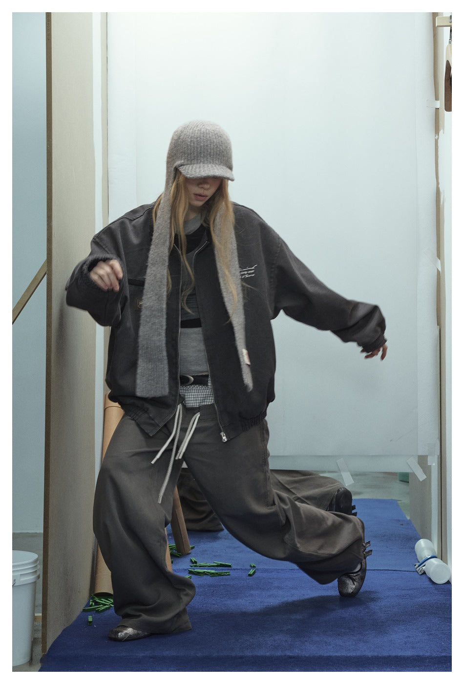 Model wearing an oversized charcoal zip jacket with a gray knit cap and scarf, and loose dark trousers.