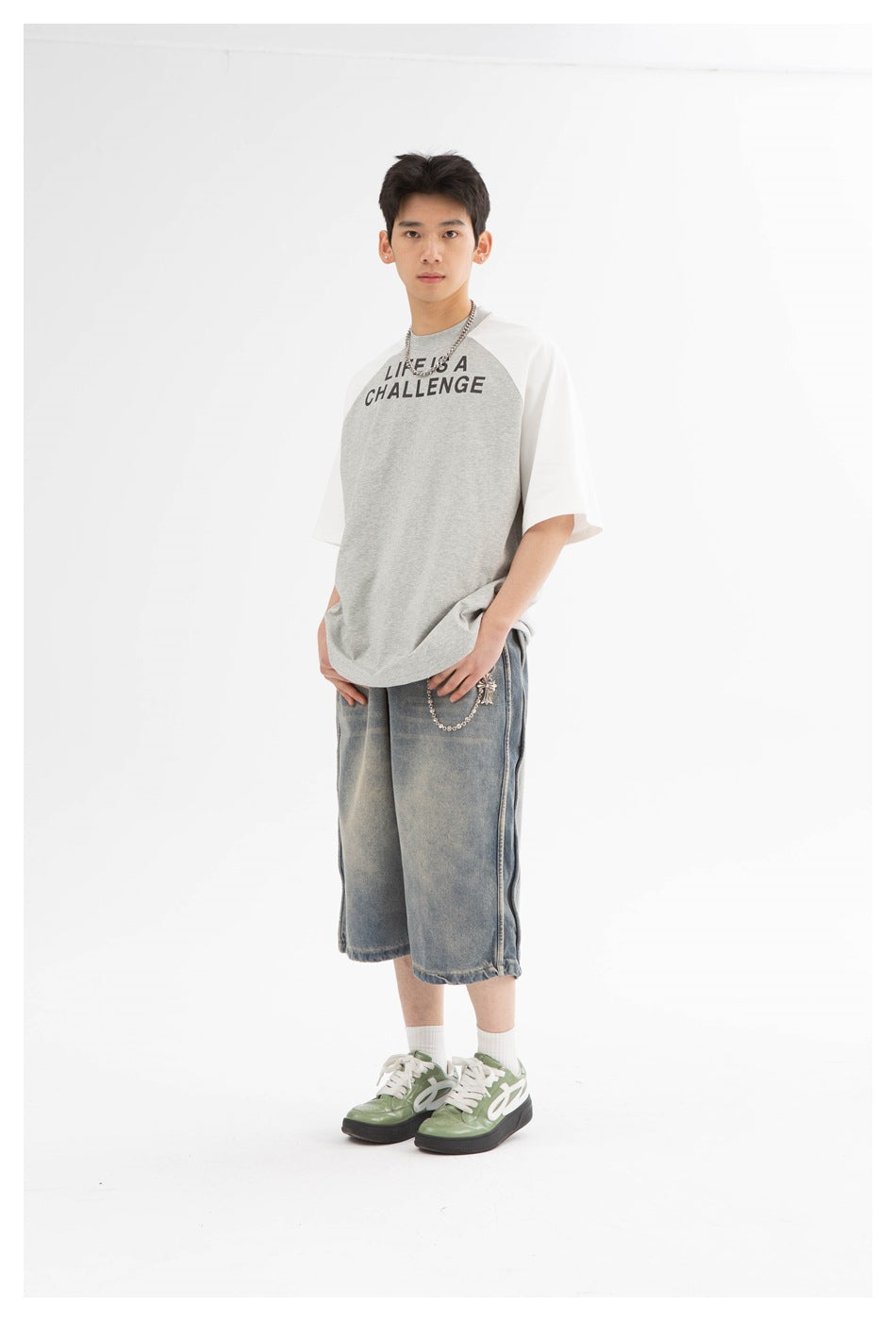 Model wearing a gray-and-white raglan T-shirt with black “LIFE IS A CHALLENGE” text and loose denim shorts.