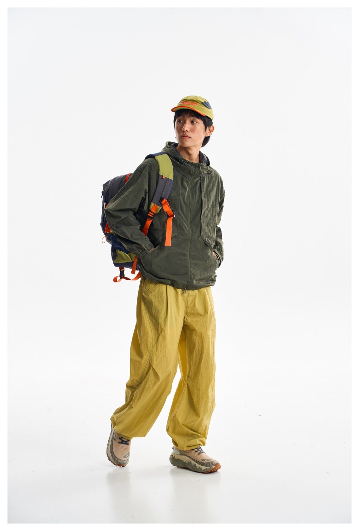 Model wearing a dark olive hooded jacket and loose mustard-yellow trousers, posed at an angle with a backpack.