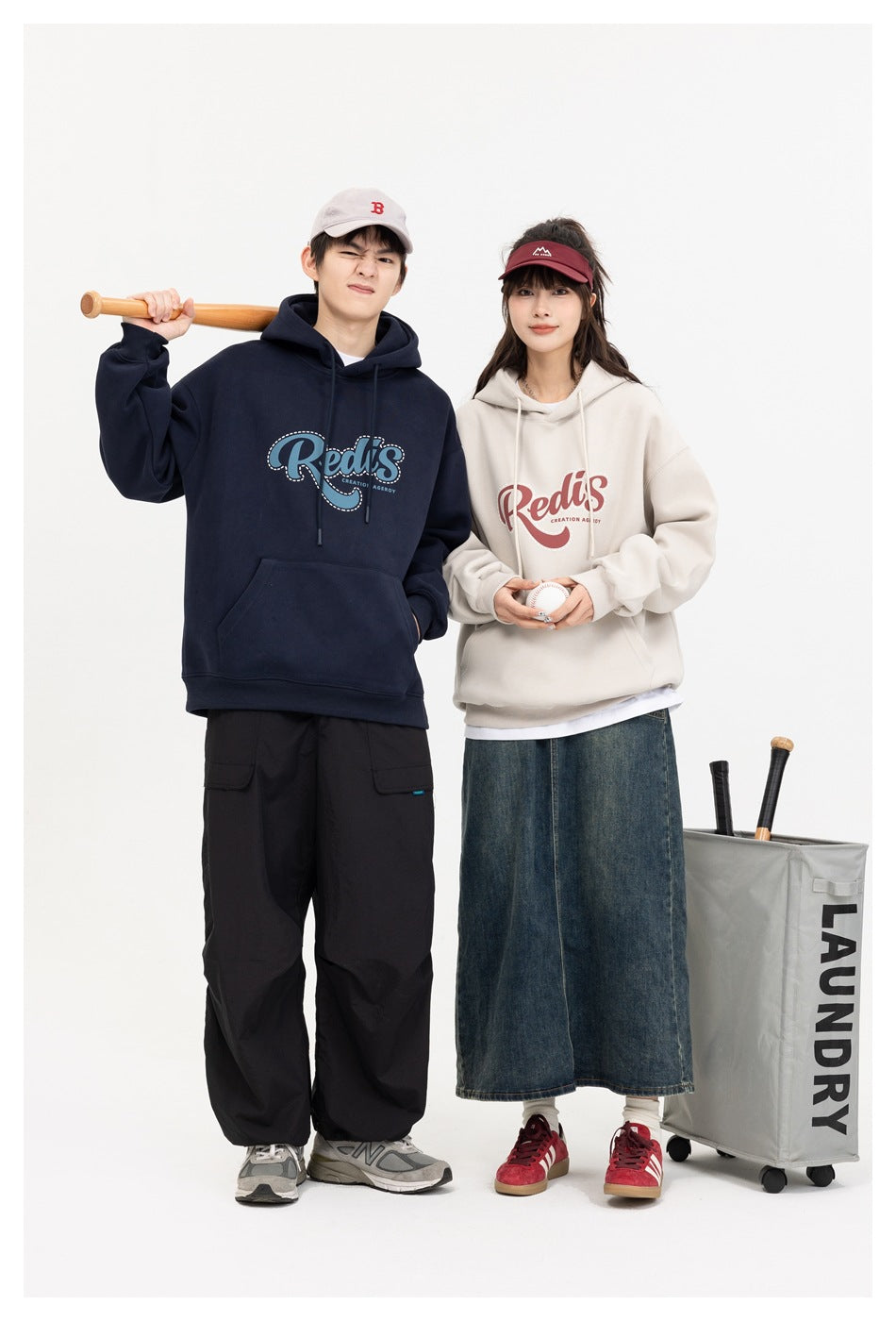 Two models wear navy and cream graphic hoodies; one holds a bat and the other a baseball.