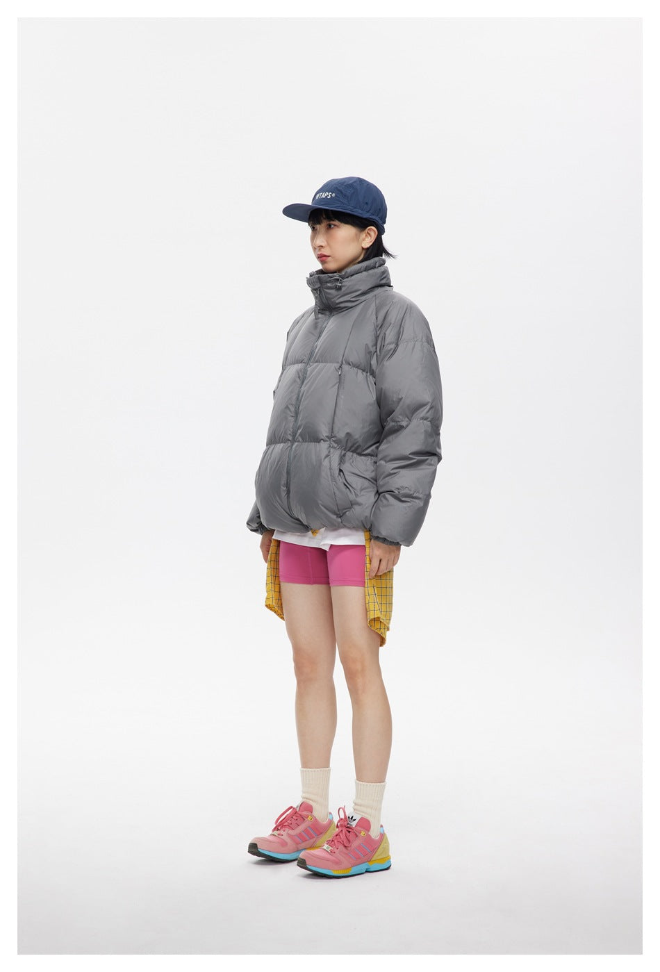 Side view of a model wearing a gray puffer jacket with pink shorts and a yellow checked shirt tied at the waist.