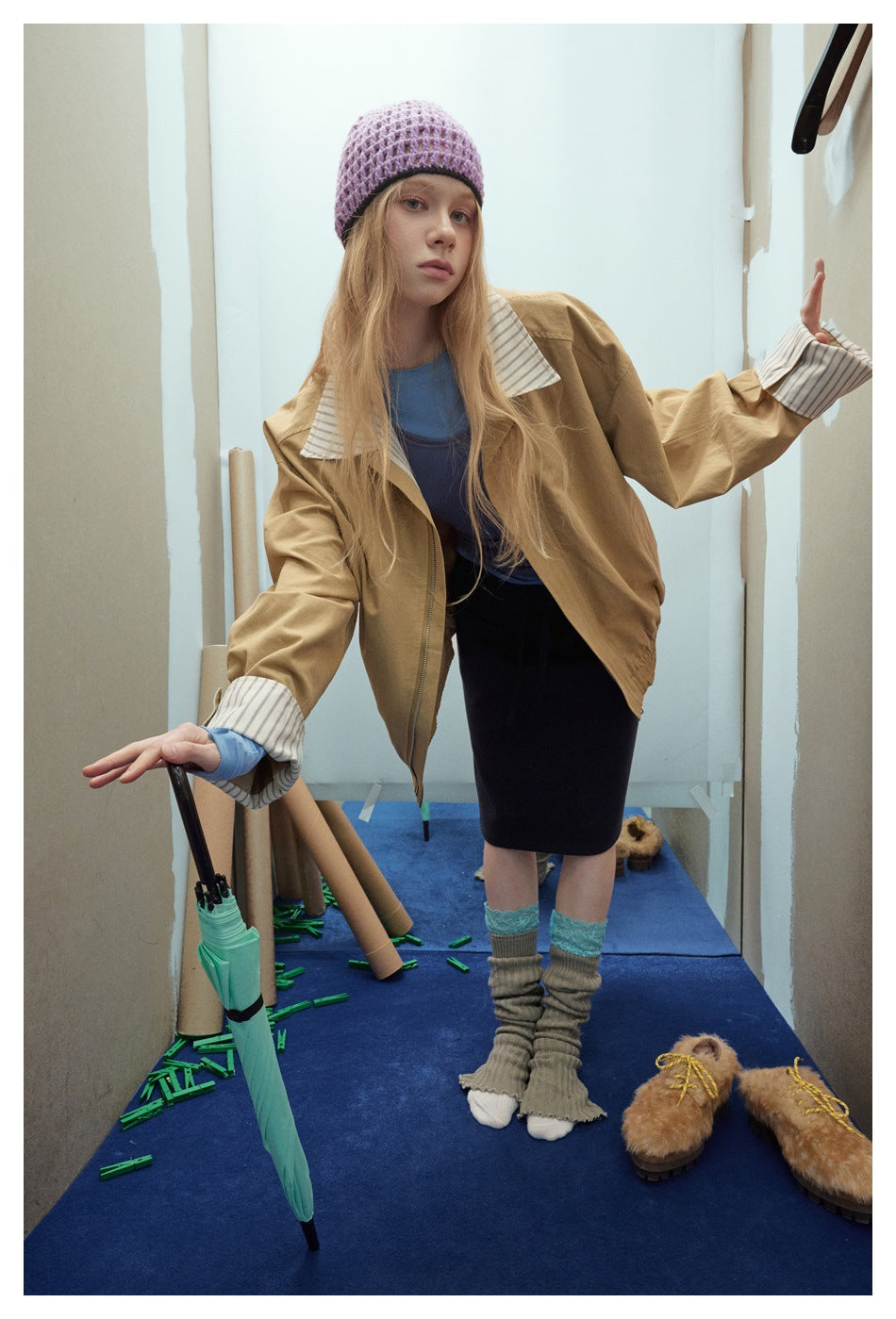 Model in a tan jacket with striped collar and cuffs, blue layers and a dark skirt, leaning beside a mint umbrella.