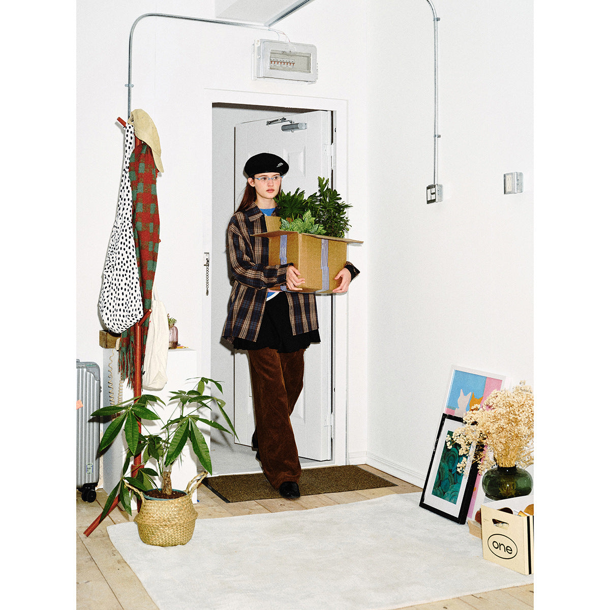 Model wearing a blue, brown and beige plaid shirt and brown wide-leg pants while carrying a box of plants.