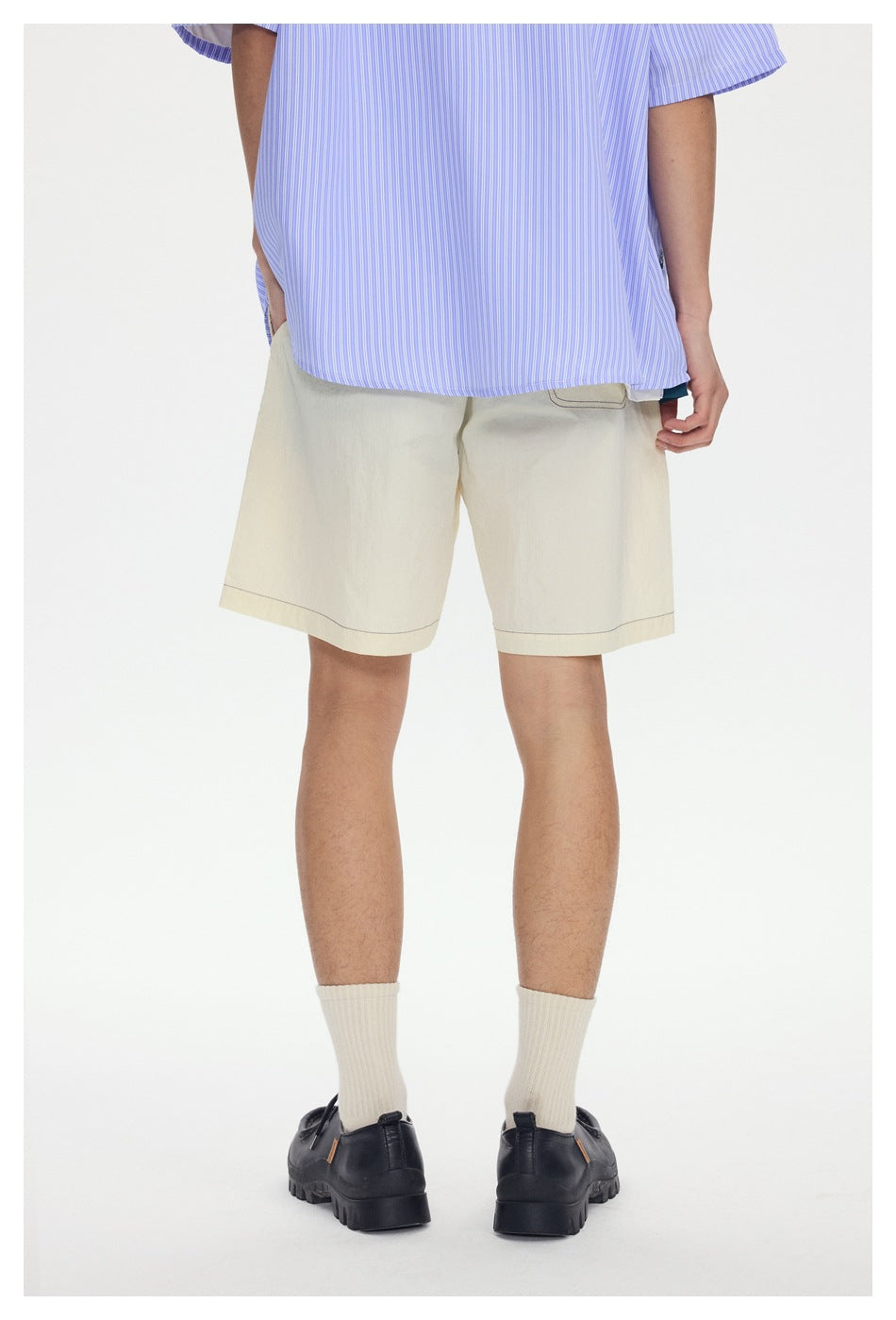 Rear view of a model wearing cream knee-length shorts with contrast stitching and a blue striped shirt.
