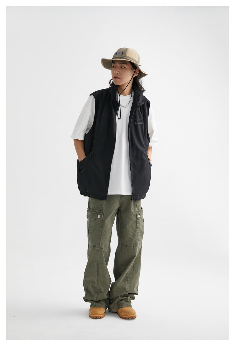 Model wearing the black zip vest open over a white tee with olive cargo pants, bucket hat and boots, full-length view