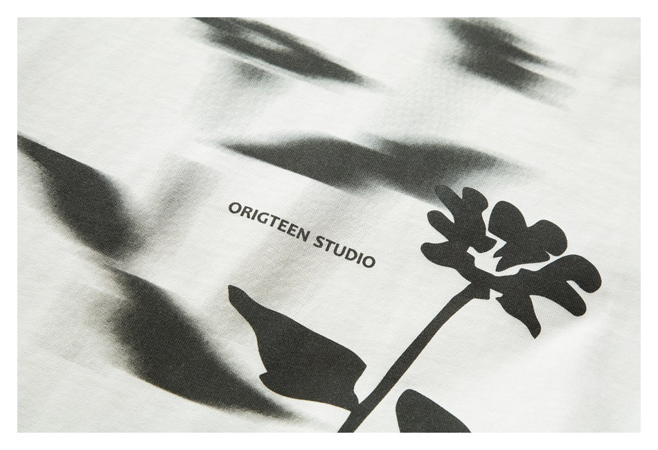 Close-up of a white shirt with a black flower graphic, blurred print, and the words “ORIGTEEN STUDIO.”
