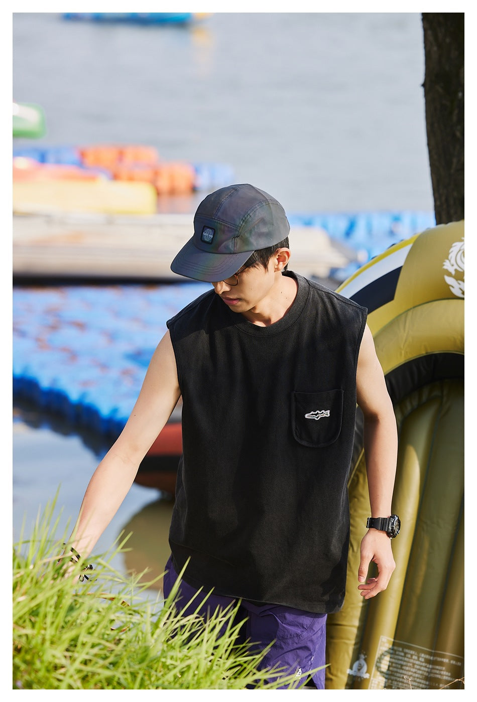 Model wearing a black sleeveless shirt with a chest pocket and purple shorts, shown outdoors.