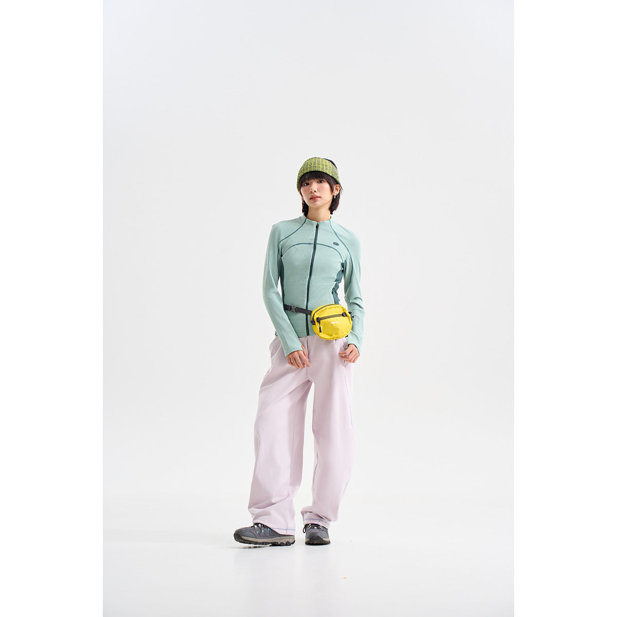 Model wearing a mint-green zip-front jacket with dark teal panels, pale lavender trousers, a knit headband and yellow waist