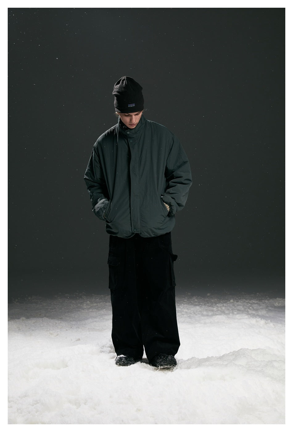Full-length view of a model wearing a dark charcoal jacket, black beanie and loose black cargo pants.