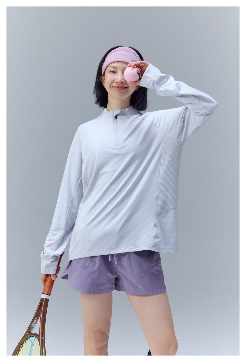 Model wearing a pale gray quarter-zip top and lavender shorts, holding a pink ball near her face.