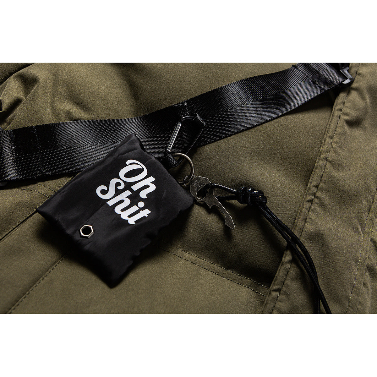 Close-up of an olive jacket’s black crossbody strap, clip, cord, and attached pouch.