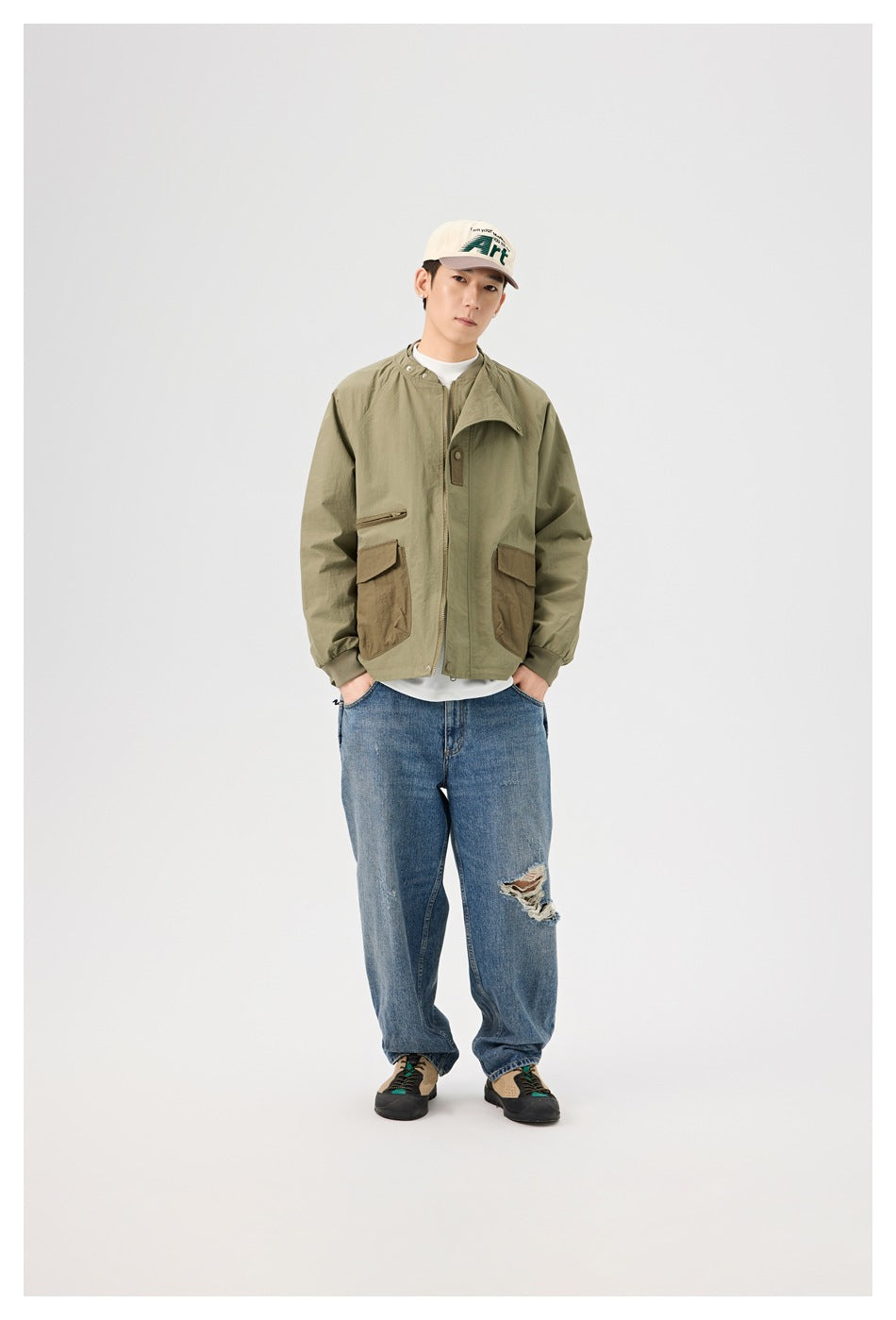 Full-length view of a man wearing an olive jacket with brown flap pockets, blue jeans, and a cream cap.