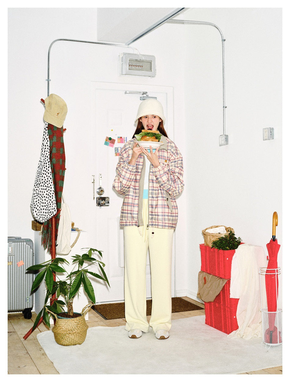 Model wearing a pastel plaid zip jacket and cream wide-leg pants, holding a sandwich in a bright entryway.
