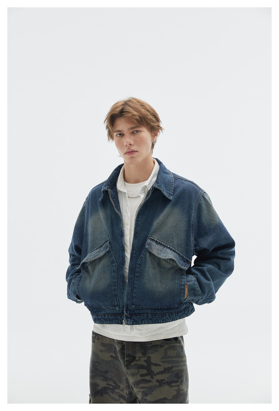 Model wearing a dark blue faded denim jacket with a zip front and flap pockets, layered over white tops and camouflage pants.