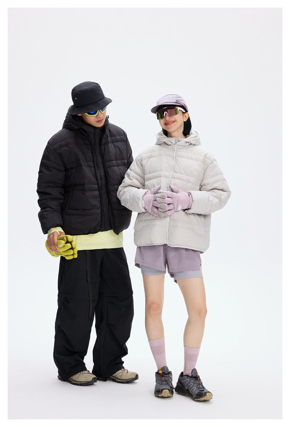 Two models wear black and light gray quilted hooded jackets; the woman holds her hands together in lilac gloves.