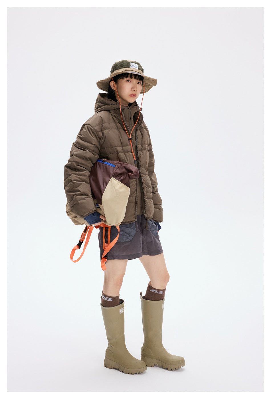Three-quarter full-length view of a model wearing a brown hooded puffer jacket and gray shorts.