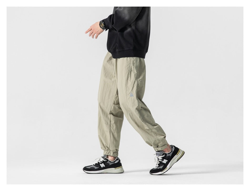 Side view of a model wearing light khaki loose jogger pants with elastic cuffs and a small blue emblem.