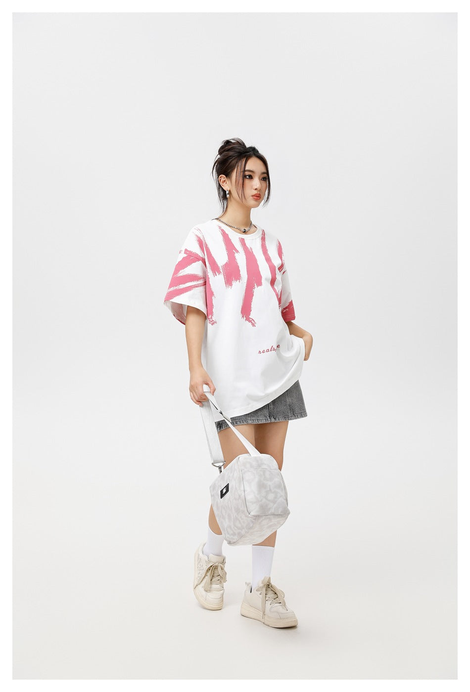 Model wearing an oversized white T-shirt with pink brushstroke graphics and small pink lettering, paired with a gray skirt.