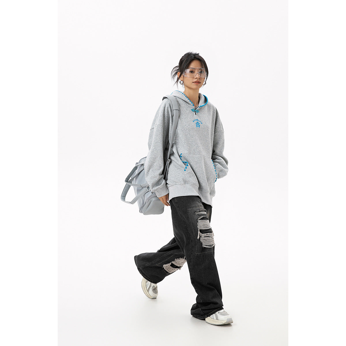 Model walking in an oversized gray hoodie with blue trim and black distressed jeans, carrying a gray bag.