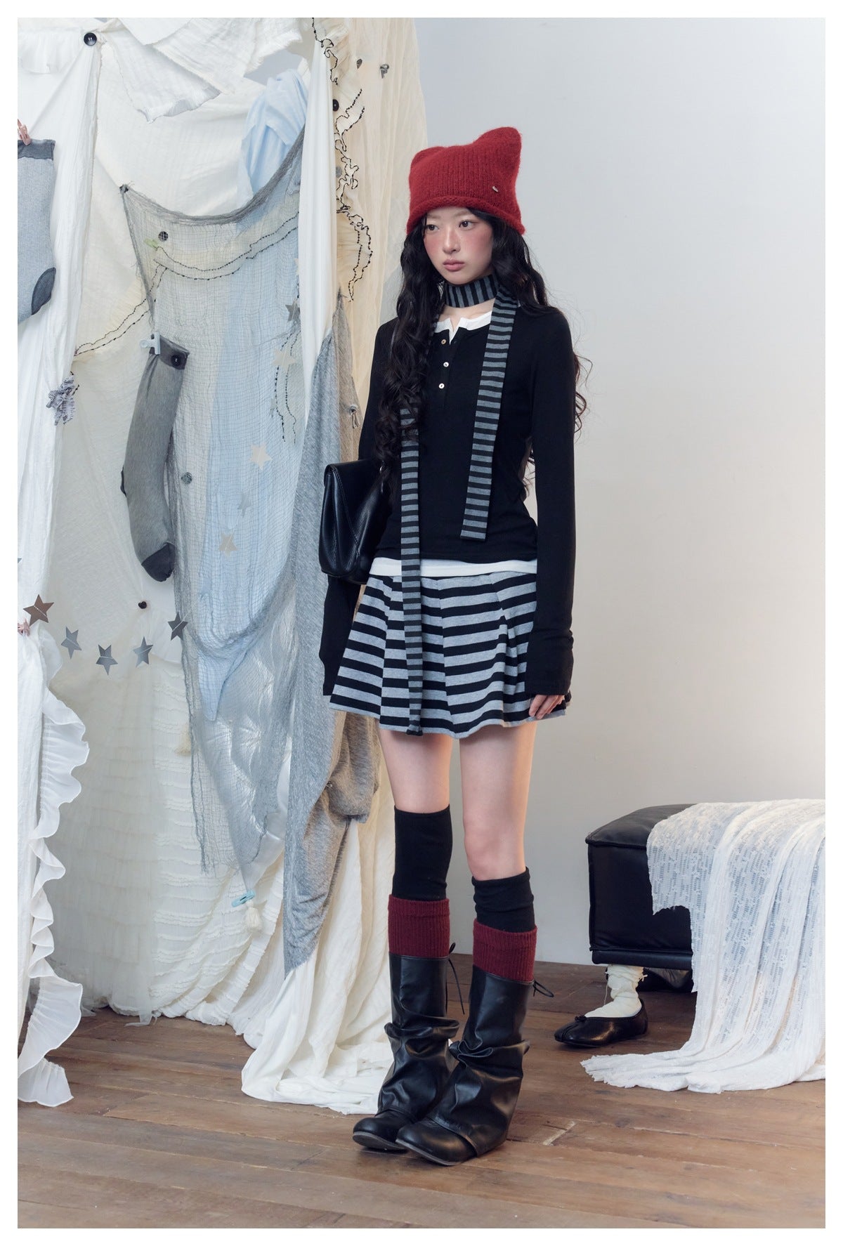 Model wears a black long-sleeve top with a gray-and-black striped mini skirt and scarf, red hat, and tall black boots.
