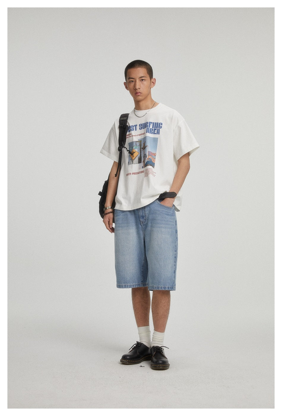 Model wearing a white graphic T-shirt and light blue denim shorts, with a black shoulder bag.