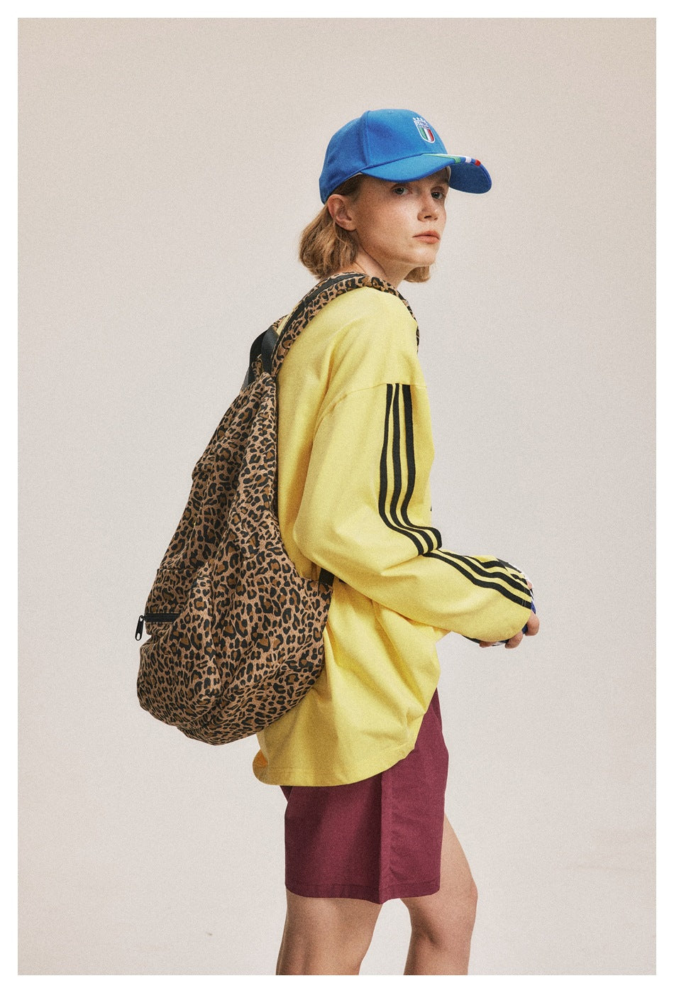Side view of a model in a yellow striped-sleeve shirt and burgundy shorts, carrying a leopard-print backpack.
