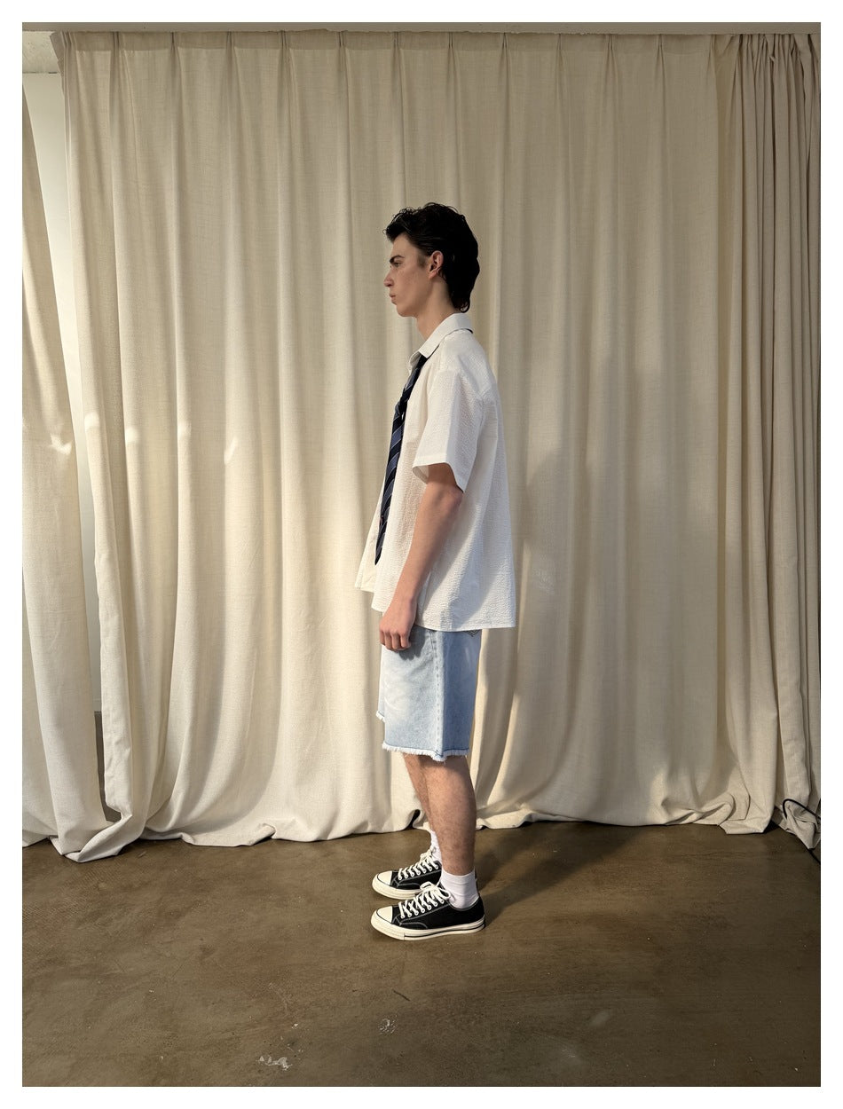 Side view of a model wearing a white short-sleeve collared shirt, striped tie and light denim shorts.