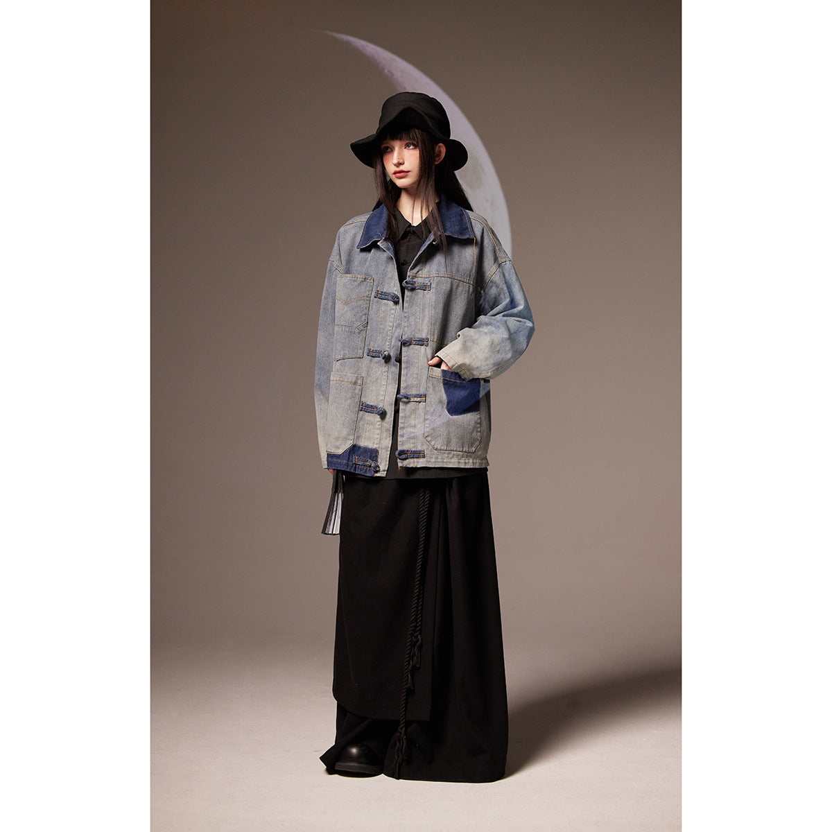 Model wearing an oversized gray denim jacket with dark blue panels and frog closures over a long black outfit.